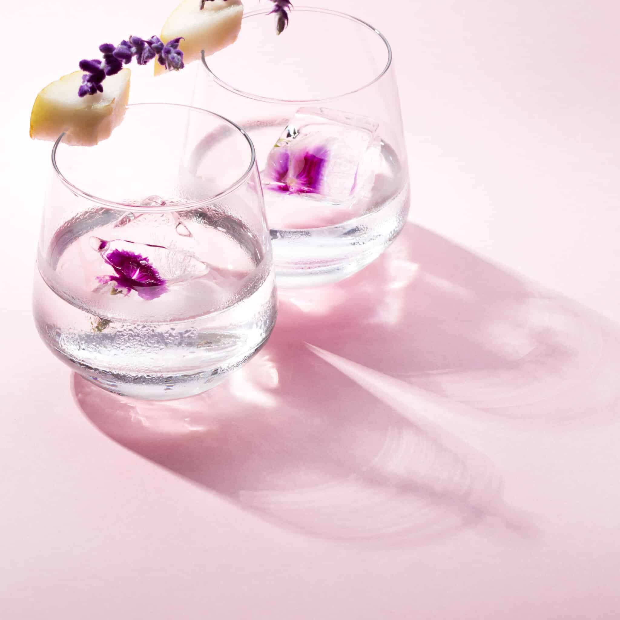 Gin cocktail on pink backdrop with shadow pear and lavender garnish and ice cube with edible flowers