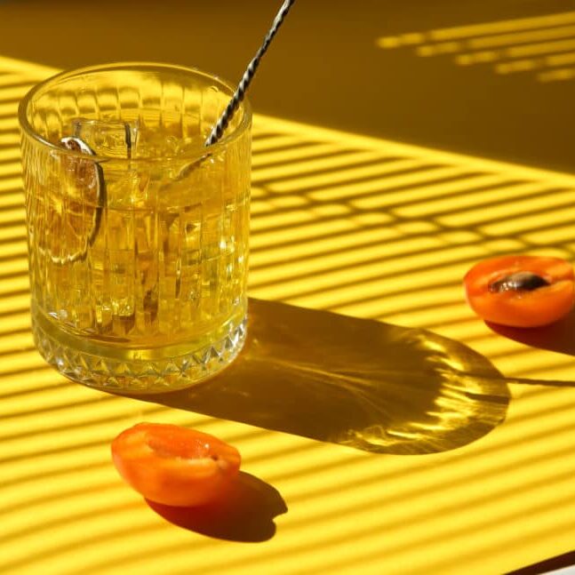 Drinks and cocktails. Minimalism. Whiskey with ice in a clear glass with a bartender's spoon, lime chips and fresh apricot on a yellow background. Sunny bright light. Background image, copy space